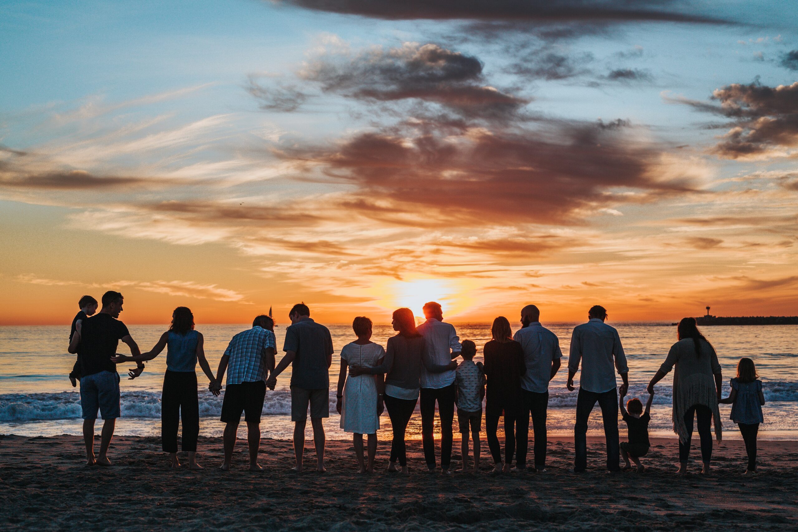 group of people holding hands and facing the ocean on a beach during sunset